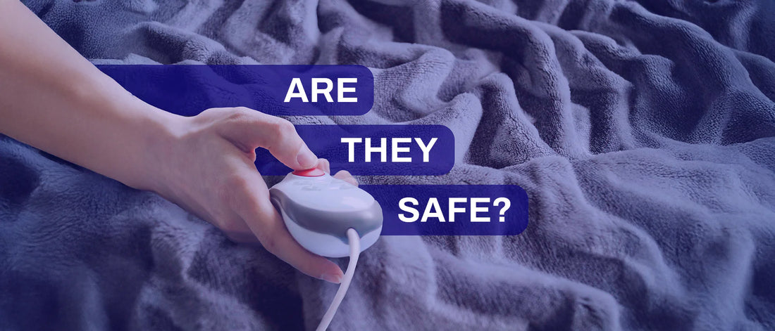 A close-up of a person's hand holding a wired controller with a red button, likely for an electric blanket, against a soft, plush gray fabric background. The text overlay reads, 'ARE THEY SAFE?' in bold white letters on a dark blue background.