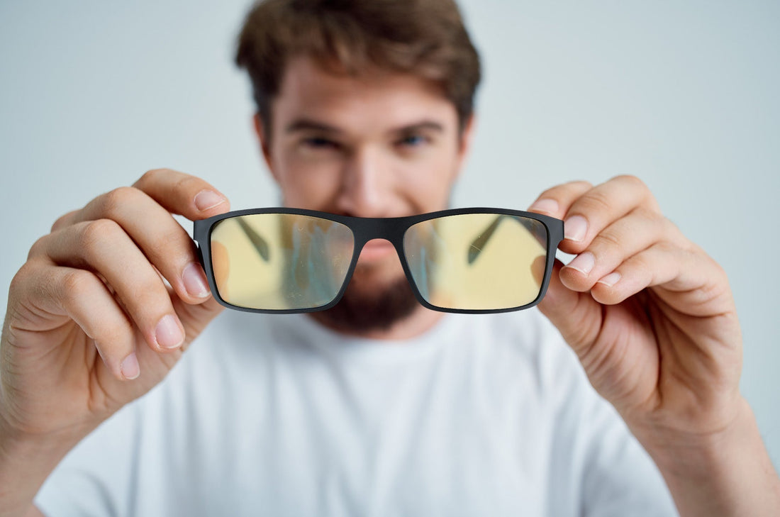 Man holding yellow-tinted blue light blocking glasses in front of the camera, focusing on the glasses while the person in the background is blurred.