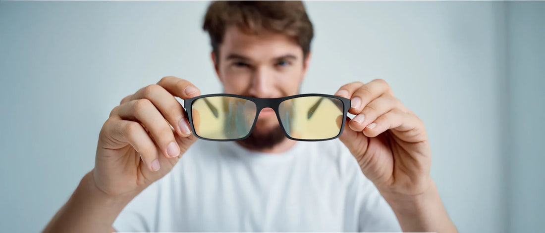 Man holding yellow-tinted blue light blocking glasses in front of the camera, focusing on the glasses while the person in the background is blurred.