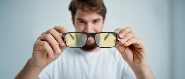 Man holding yellow-tinted blue light blocking glasses in front of the camera, focusing on the glasses while the person in the background is blurred.