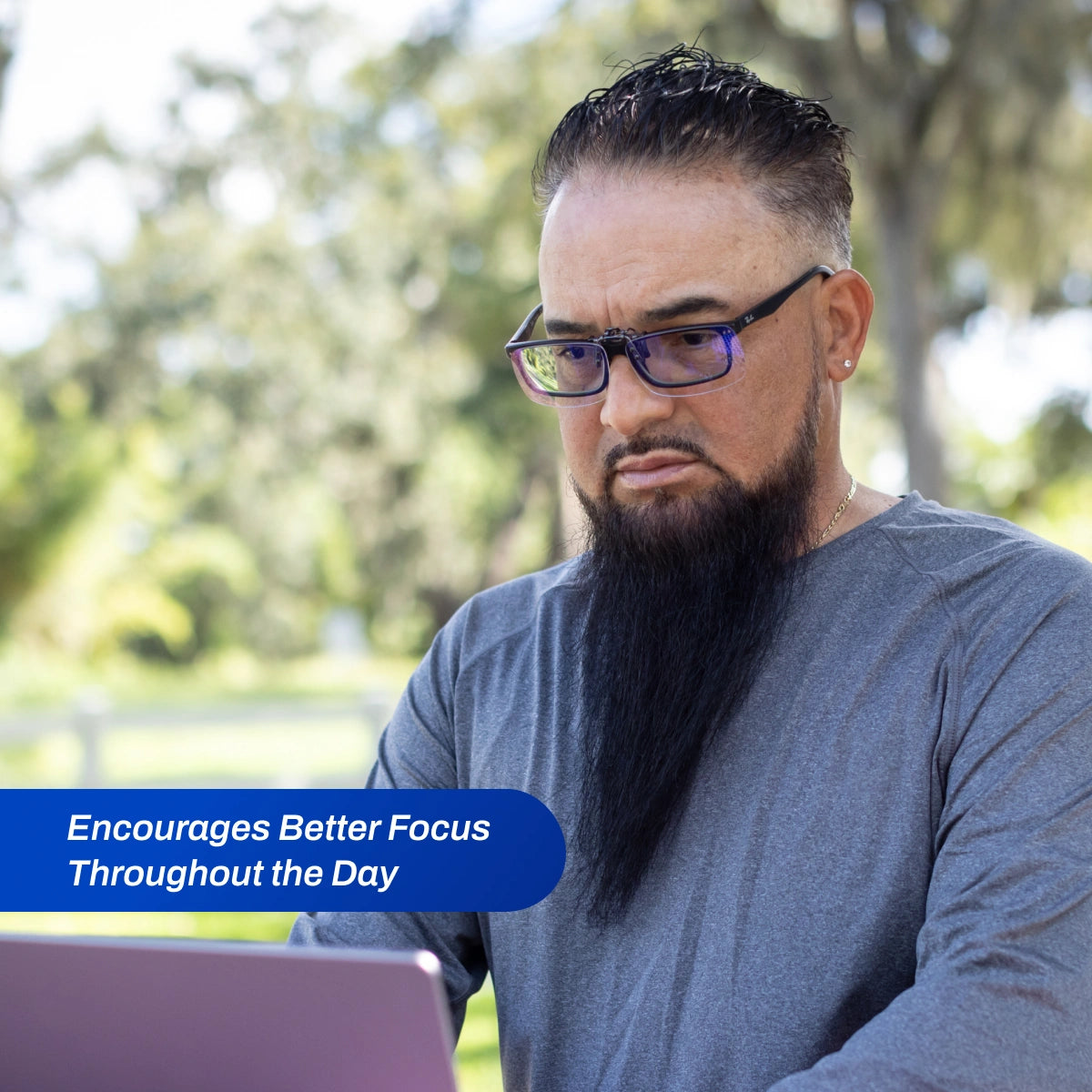 Man with Clip-ons Blue Light Glasses and a beard using a laptop outdoors with text 'Encourages Better Focus Throughout the Day'.